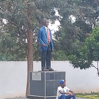 An outdoor scene in Machakos Central ward, Kenya, prominently features a full-body statue of an adult male positioned centrally. The statue depicts a dark-skinned individual with short dark hair, dressed in a blue suit jacket, white shirt, red tie, dark trousers, and dark shoes. It stands on a multi-tiered, dark gray rectangular pedestal, which has a rectangular, light-colored informational plaque affixed to its front. To the right of the pedestal, a young individual, presumed male, is seated on the ground. This person wears a white t-shirt with a graphic design, blue jeans, and a multi-colored baseball cap worn backwards, facing left towards the statue. The background consists of dense green foliage from various trees and a horizontal light-colored wall. The ground in the foreground is a light brown, earthy surface.