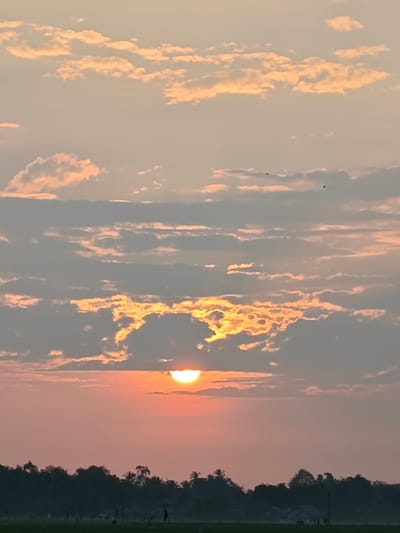 The image displays an outdoor scene in the city of Mee Pangwa, Indonesia, depicting either a sunrise or sunset over a flat horizon. The upper 75% of the frame is dominated by a sky filled with multiple cloud layers. The sun is visible as a bright, luminous orange-yellow semi-circle, positioned low in the sky and partially obscured by a dense, dark cloud band directly above it. Sunlight illuminates the undersides and edges of various clouds, creating orange and yellow highlights amidst the predominantly grey and pale orange cloud formations. Two small, dark, indistinct specks, possibly birds or distant airborne objects, are present in the upper-mid section of the sky. The lower 25% of the image shows a dark, silhouetted landscape. This silhouette consists of a dense line of tropical vegetation, including discernible palm trees, with a flat expanse of ground, possibly agricultural fields, extending to the horizon. Faint, indistinct human figures and small, low-profile structures are minimally discernible on the distant horizon within the dark silhouette, towards the right side of the frame. A slender, dark, vertical object, resembling a pole or stake, is observed in the lower-middle foreground within the silhouetted landscape. No explicit actions or interactions are discernible among the indistinct figures.