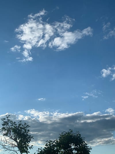 This is a photograph of the sky with some trees in the foreground, taken in Карманово, Russia. The sky is a clear blue with scattered white clouds. The clouds in the upper portion of the image are wispy and light, while a band of darker, more substantial clouds stretches across the horizon in the lower part of the image. On the left and right sides of the frame, the tops of trees with green foliage are visible. The overall scene depicts a natural outdoor environment on a day with varied cloud cover.