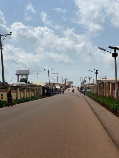 The image depicts a street scene in Tangu, Nigeria. The street is paved with reddish-brown asphalt and is lined with buildings, shops, and vegetation. Utility poles with wires and streetlights are prominent along both sides of the road. A person is walking away from the camera on the left side of the street, wearing dark pants and a light-colored shirt with a backpack. Several other individuals and motorcycles are visible further down the street, suggesting moderate activity. The sky is partly cloudy. In the background, a large water tower stands behind a fence. The overall impression is of a typical urban or semi-urban environment in Nigeria.