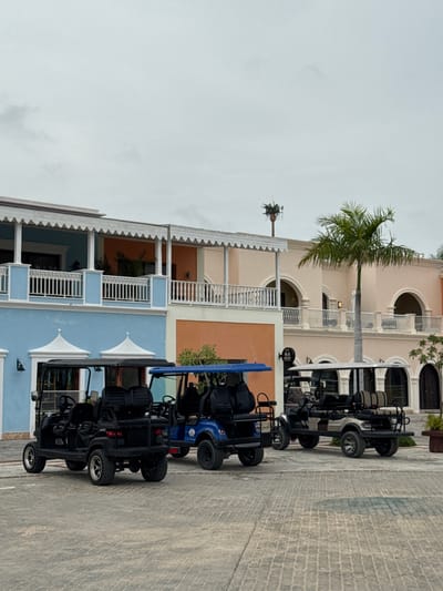 In Higüey, Dominican Republic, under a cloudy sky, four golf carts are parked on a paved courtyard in front of a multi-story building with pastel-colored facades. The building features white railings, arched doorways, and balconies. A palm tree stands to the right of the building. The golf carts, one blue and three black or gray, are unoccupied. The scene is serene and depicts an outdoor area likely associated with a resort or residential complex.