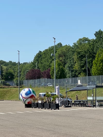 The image shows an outdoor parking lot in Uffenheim, Germany, with a large, inflated soccer ball as the central focus. The ball, which is white with multi-colored geometric patterns in blue, green, and red, is propped on black planters. To the right of the ball, there are more black planters containing small palm trees. Behind the planters, a white van is partially visible, and further back, a fence separates the parking lot from a grassy hill covered with trees. Several streetlights are visible throughout the scene, along with a black patio umbrella and a seating area with picnic tables and benches. A person, whose gender is not clearly identifiable, is seated at one of the tables. The sky is clear and blue.