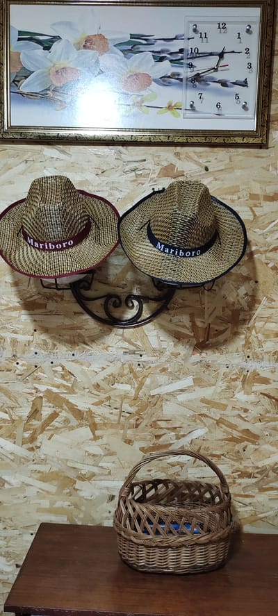 The image displays an interior scene, likely a domestic setting, featuring two straw hats hanging on a decorative metal hook mounted on a wood-paneled wall. Both hats have a band with the word "Mariboro" embroidered on them. Above the hats, a framed picture of daffodils and pussy willows hangs on the same wall, with a clock integrated into the picture frame. Below the hats, a wicker basket sits on a wooden table. The overall impression is one of rustic decoration.
The location of Dimitrovgrad, Russia, is not determinable from the visual information provided in the image.