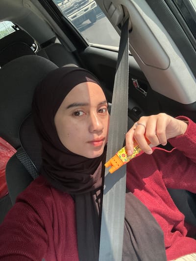 The image depicts a single individual, likely female, seated in the front passenger seat of a vehicle. She is wearing a dark brown hijab and a dark reddish-brown ribbed cardigan over a dark inner garment. A grey seatbelt is fastened diagonally across her chest. Her right hand, adorned with a silver ring on the ring finger, holds an orange and yellow dispenser-like object, possibly a candy dispenser, featuring fruit graphics and partial text "Juice." Her face is partially illuminated by direct sunlight from the right, casting a shadow across her left side. In the background, visible through the car window, is a white commercial truck, possibly with "SAMATRA" (or similar partially visible text) on its grille and a "B" prefix on its license plate, suggesting an Indonesian registration. The vehicle appears to be on a street or road during daylight hours. This scene is situated in the city of Entalsewu, Indonesia.