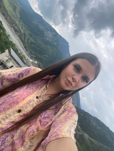 A woman with long, dark hair and heavy eyeliner is taking a selfie outdoors, with a mountainous landscape in the background. She is wearing a colorful, patterned shirt. The image appears to be taken from a high vantage point overlooking a valley with a river or road, and some construction in the distance. The sky is cloudy. The location is specified as Chalda, Russia.