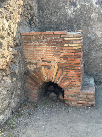 The image shows an ancient brick oven in Pompeii, Italy. The oven is made of red and yellow bricks, with a semicircular opening at the bottom. The surrounding walls are made of rough stone and concrete. The ground in front of the oven is covered with gravel and small plants. The scene appears to be well-preserved, suggesting it is part of a historical site open to the public.