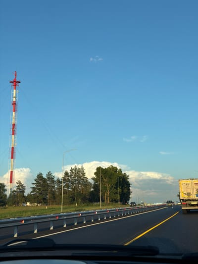 The image depicts a view from a vehicle traveling on a highway in Egor'evo, Russia. In the distance, a tall, red and white striped radio tower stands against a clear blue sky with scattered clouds. The highway is paved and features a yellow dividing line. A metal guardrail with reflective orange markers separates the lanes. Several cars and a truck are visible on the road, moving in the same direction. The surrounding environment includes a grassy field with trees and power lines. The lighting suggests it is either early morning or late afternoon.