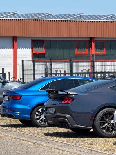 A blue sports car is parked alongside a gray sports car. The blue car has a rear spoiler and distinct taillights. A German license plate reads "OG MW 666". Behind the cars, a building has a brown-paneled roof, white walls, and green paneling with red-framed windows. A metallic structure with wires and bars is visible between the cars and the building. Solar panels are affixed to the roof. The pavement and a grassy verge are on the right.