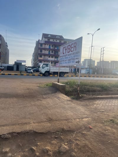 This media file captures a bright, sunny street scene in Gitothua ward, Kenya, likely during the morning or late afternoon. The main subject is a prominent directional sign for a "CYBER" cafe, made of white material with red text, featuring an arrow pointing to the right. The sign is positioned at the edge of a dirt and gravel path that runs alongside a paved road.
The services listed on the sign are:
* CYBER
* PRINTING
* PHOTOCOPY
* BUSINESS CARD BINDING
* COMPUTER TRAINING
* KRA PIN & REGISTRATION
* FILING RETURNS
* WI-FI SUBSCRIPTION (20 Mbps)
* RENTING OFFICE SPACE
* PASSPORT PHOTO
In the background, multiple buildings of varying heights and colors are visible, including a multi-story structure with purple and beige facades. Utility poles with numerous overhead wires extend across the sky. Several vehicles, including a white truck and other cars, are parked or driving on the paved road, indicating active traffic or commerce. The immediate foreground shows a rough, unpaved area with some scattered debris and sparse vegetation. The clear sky suggests fair weather.