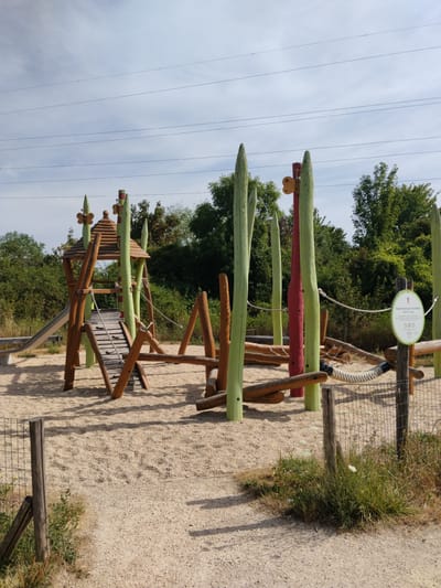 The image shows a natural playground constructed with wooden elements and tall, green, painted poles resembling reeds. In the center is a multi-level wooden structure with a roof, a slide, a ramp with wooden steps, and various other climbing and balancing features. Ropes are integrated into the playground's design, serving as railings or climbing aids. The ground is covered with fine sand or gravel.
To the right, a circular sign with a pictogram of a child and text in French is mounted on a wooden post. The sign indicates that the area is a play space for children aged 5 to 12 years old and displays several pictograms related to safety rules, though the details are not legible.
In the background, there is lush green vegetation consisting of trees and bushes. The sky above is partly cloudy, and several horizontal power lines are visible in the upper portion of the image. The overall scene depicts an outdoor recreational area designed to blend with a natural environment. This playground is located in the Parc de l'Hôtel de Ville in Rosny-sous-Bois, France.