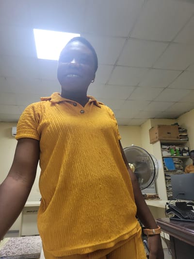 The image shows a medium shot of a woman wearing a bright yellow, textured polo shirt, standing in what appears to be an office environment. She has dark skin and short, dark hair, and is looking towards the camera with a slight smile. She is wearing a light brown wristband. The background shows a fluorescent light panel in the ceiling, a desk with a computer monitor and a landline telephone, a standing fan, and shelves filled with papers and boxes. The overall lighting suggests the photo was taken indoors during the day. The specific location in Abuja, Nigeria, cannot be determined from the visual information provided.