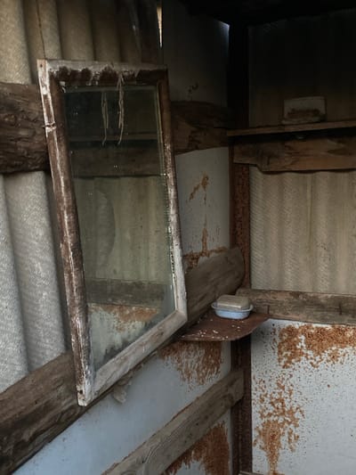 The image depicts the interior of a rustic, possibly abandoned structure in Cairaclia, Moldova. A weathered, wooden-framed mirror, tilted at an angle, is the central object. Its surface is dusty and reflects a dimly lit, corrugated wall and wooden beams. To the right of the mirror, a small, rusty shelf holds a bar of soap in a pale blue dish. The walls are a mix of corrugated material and what appears to be painted metal, heavily stained with rust. Wooden beams are integrated into the structure, supporting shelves and adding to the aged, disheveled aesthetic. The lighting is low, suggesting an interior space with limited natural light.