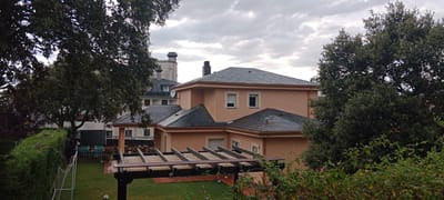 This is a medium-distance, eye-level exterior shot taken on an overcast day in Los Ángeles de San Rafael, Spain. The scene depicts a residential backyard with a prominent two-story house in the center, painted a light peachy-orange color. The house has a dark gray slate roof with several gabled sections and dormer windows. A wooden pergola structure with a slatted roof is situated in the foreground, partially obscuring the lawn. The lawn is green and appears damp, with a soccer ball visible on the right side. To the left of the house, a dark green hedge runs along a white metal soccer goal. Behind the house, a larger, multi-story building with a light-colored facade and a tall chimney is visible, along with another building with a dark roof. Mature trees with dense green foliage frame the scene on the left and right sides, their leaves slightly blurred by the wind. The sky is filled with gray, cloudy cover.