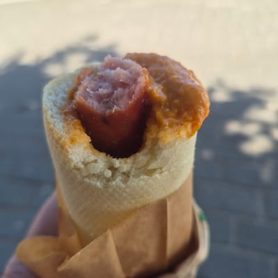 A close-up, slightly overhead shot shows a hot dog wrapped in brown paper. The hot dog is mostly obscured by a bun and a thick, orange sauce. The visible portion of the hot dog is a pale pink with a slightly rough texture, suggesting it might be a sausage or frankfurter. The bun is soft and white, with a torn opening revealing the hot dog. The sauce appears to be a tomato-based sauce, possibly with spices, and has a glossy sheen.

The background is softly blurred, showing a pattern of light and dark grey shapes that could be shadows cast by trees or other objects on a paved surface. The lighting suggests it is daytime, with the shadows indicating sunlight. The overall impression is of a casual, outdoor setting. The image is cropped tightly on the hot dog, with no clear indication of people or a specific location beyond what can be inferred from the background. The dominant colors are the beige of the bun and paper, the orange of the sauce, and the muted tones of the background. There is no visible text in the image.