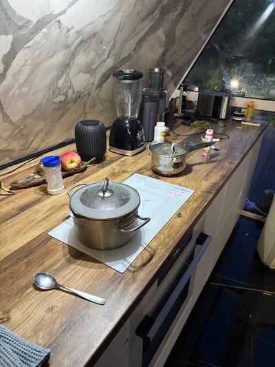 In the dimly lit kitchen of a residence in Vol'noe, Russia, a wooden countertop displays an assortment of kitchenware and appliances. A lidded pot sits atop a white induction hob, with a small spoon resting to its left. Nearby, an apple rests on a decorative leaf next to a shaker bottle. To the right, a blender and a toaster are visible, with other miscellaneous items scattered across the counter. The kitchen features a marble-patterned backsplash and a window that offers a glimpse of the outdoors at dusk. The floor is tiled in a dark, reflective material. The scene suggests a domestic environment where cooking or food preparation is underway.