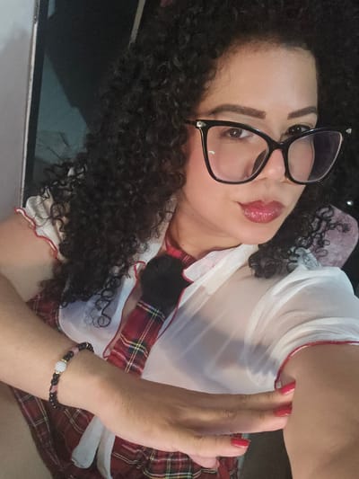 A woman with curly hair and glasses is lying down in a vehicle. She is wearing a white, sheer blouse with red trim over a plaid top and tie. She has a beaded bracelet on her wrist and red nail polish. The location is unknown.