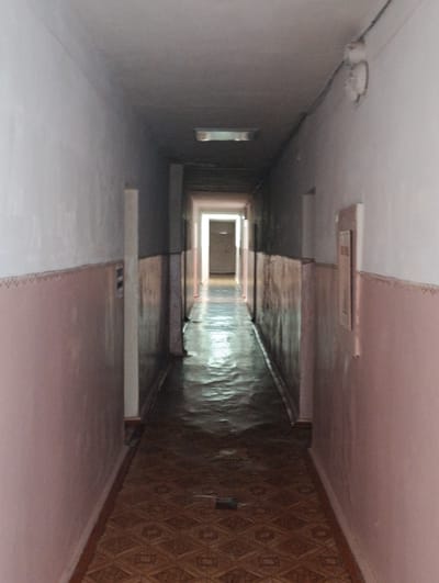 The image shows a long, dimly lit hallway in a building, possibly a dormitory or institutional facility. The hallway has pale pink walls with a decorative border near the floor. The floor is tiled with a patterned linoleum. A light fixture is visible on the ceiling. The hallway recedes into the distance, where a bright light emanates from an open doorway. There are no people visible in the image. The image appears to be taken in Khorol, Ukraine.