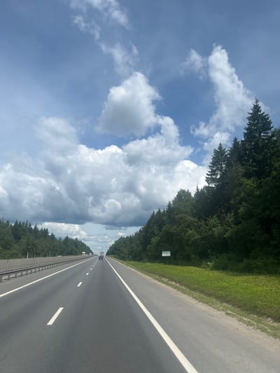 This media file depicts a road scene with a dramatic sky. The road, a dark asphalt surface, has a clear white line marking the lanes and extends into the distance, suggesting a highway. The edge of the road is bordered by lush green grass and dense, dark green coniferous trees that line the route on both sides, creating a tunnel-like effect. The sky is a prominent feature, filled with large, fluffy white clouds against a bright blue backdrop. Some of the clouds appear to be gathering, forming a dramatic, almost storm-like formation in the center of the frame. A few vehicles are visible in the distance on the road, appearing as small shapes. The overall scene suggests daytime, possibly mid-afternoon, given the brightness of the sky and the shadows cast. There is a small, rectangular sign visible on the right side of the road, amidst the trees, with some text on it. While the text is difficult to read, it appears to be green with white lettering. The image conveys a sense of travel and perhaps anticipation, given the vastness of the sky and the open road. The location is identified as Komfort, Russia.