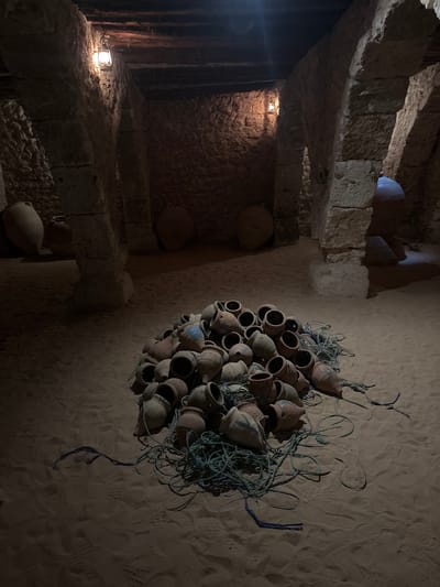 In the city of Tezdaine, Tunisia, an interior scene is depicted within a dimly lit, rustic structure constructed of rough-hewn stone and wood beams. The floor is covered in sand, with distinct footprints visible. In the foreground, a large pile of clay pots of various sizes is gathered, some exhibiting blue painted markings. These pots are entangled with lengths of blue and green rope. In the background, larger, smooth, light-colored amphorae are positioned against the stone walls. Two old-fashioned oil lamps, suspended from the ceiling, cast a warm, localized light, illuminating the stone arches and the rough texture of the walls. The overall atmosphere is one of historical storage or display.