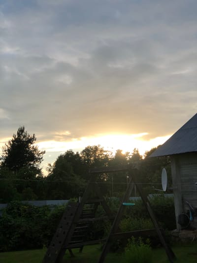 The image shows a backyard scene at dusk. The sky is filled with overcast clouds, with a bright band of sunlight peeking through the clouds on the horizon. In the foreground, a wooden swing set is visible, along with lush green vegetation. To the right, a portion of a wooden building with a corrugated metal roof is visible, and a satellite dish is mounted on its side. A tall pine tree stands to the left, silhouetted against the sky. The overall impression is one of a rural or suburban setting in Zhukovo, Russia, at the end of the day.