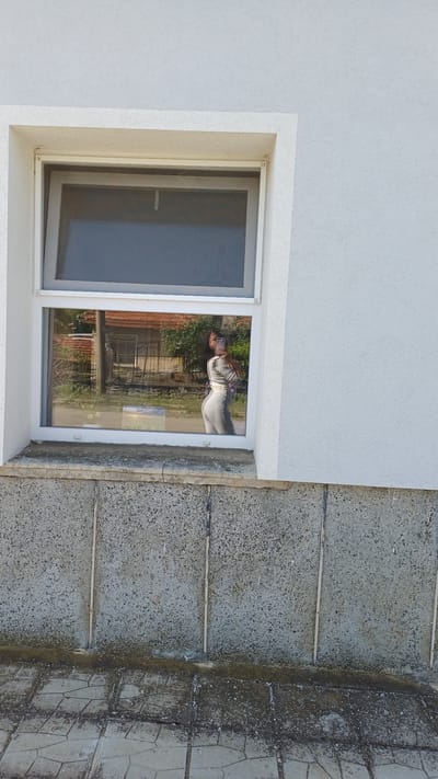 A low-angle shot depicts the corner of a building. The building's wall, featuring a rough, white texture, meets a gray, multi-textured exterior ground surface or sidewalk. A window is set into the wall; a partially open screen is visible on the left, and a reflection in the glass pane shows a person holding a camera or phone and an outdoor scene with a building with a red roof. The ground or sidewalk is made of varied materials, including patterned tiles and what appears to be poured concrete with separating strips. The location is Gorna Verenitsa, Bulgaria.