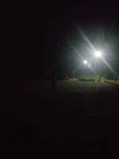 The image depicts a dimly lit outdoor scene at night, possibly a park or recreational area. Two streetlights are prominently visible, casting a bright glow and creating lens flares. The ground appears to be covered in grass and possibly some dirt paths. Trees are present in the background, partially illuminated by the streetlights. The overall impression is one of a tranquil, albeit dark, outdoor space. There is no visible indication of the specific location being in Kagoma, Nigeria.
