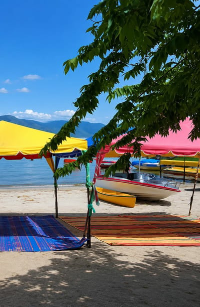 The image depicts a beach scene in Situngkir, Indonesia, on a bright, sunny day with a clear blue sky and scattered white clouds. The foreground is dominated by a large tree with green leaves casting shadows on the sand. In the mid-ground, several colorful beach umbrellas, yellow and pink, provide shade. Beneath them, rolled-up mats, one blue and striped, and another with horizontal stripes of red, yellow, and orange, are laid out on the sand.

A white speedboat with a red trim is beached on the sand, positioned behind the umbrellas and mats. Next to it, a bright yellow kayak or small boat rests on the shore. Further back, several other small boats and watercraft, some yellow and some blue, are lined up along the water's edge. The water is calm and blue, meeting the sandy beach. In the background, distant mountains are visible, their slopes covered in green vegetation, under the expansive sky. The overall atmosphere is one of a serene and inviting beachside setting, likely a recreational area.