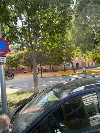 The image captures a sun-drenched urban scene in Sant Adrià de Besòs, Spain, likely during the daytime given the strong shadows. In the foreground, the roof and windshield of a dark-colored car are visible, reflecting the surrounding environment, including trees and buildings. A portion of the car's interior, featuring a steering wheel and dashboard, is also discernible through the windshield.
To the left, a blue and red circular traffic sign with a white horizontal bar is mounted on a pole, accompanied by a rectangular white sign with text that appears to read "RITZAT KHM" in black lettering. Beyond the car, a paved area resembling a sidewalk or plaza is visible, bordered by a grassy strip. Several trees with dense green foliage are scattered throughout the scene, casting dappled shadows on the ground.
In the middle ground, a group of four individuals are gathered, appearing to be engaged in a game of pétanque, with several balls on the ground. Another individual, a woman, is walking across the paved area in the background. Behind them, buildings with a predominantly pinkish-red facade are situated, with some featuring balconies. A white van is parked in front of one of the buildings. The ground in the mid-ground and background is covered with a significant amount of yellow fallen leaves, suggesting a seasonal event, likely autumn. The overall atmosphere is calm and typical of a residential urban environment on a bright day.