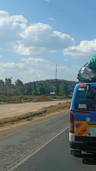 A blue and white "Toyota" bus, laden with luggage on its roof, occupies the right side of the frame, partially obscuring the view. Beyond, a dusty road and a grassy shoulder lead to a scene of sparse vegetation and a small hill. A tall transmission tower stands atop the hill, overlooking a scattering of small structures and trees. The sky is bright blue, filled with puffy white clouds. The overall impression is of a landscape somewhere rural or remote.