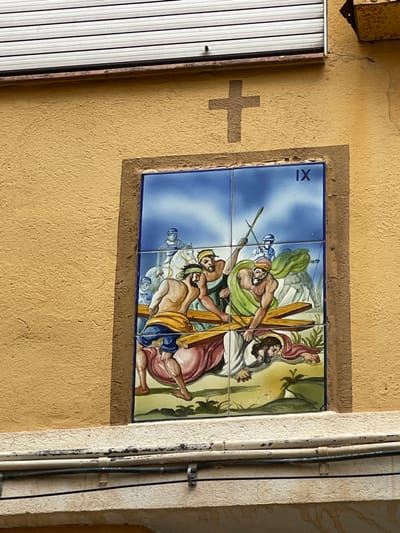 An outdoor image taken in Buñol, Spain, depicts a religious ceramic tile mosaic embedded within a textured, yellow-ochre stucco wall. The mosaic, composed of six rectangular tiles arranged in a 2x3 grid, illustrates the Ninth Station of the Cross, indicated by the Roman numeral "IX" in the top right tile.
The central scene portrays a figure resembling Jesus, clad in a reddish-pink robe, fallen on his hands and knees under the weight of a large wooden cross. Two bearded, muscular men assist him; one on the left, bare-chested with a yellow waist wrap and blue pants, lifts the horizontal beam of the cross from the left side. Another man on the right, wearing a green robe, also supports the cross, appearing to use a stick for leverage. In the background, under a blue sky, several figures are visible, including at least two men on horseback wearing blue helmets and white garments, possibly Roman soldiers. The ground below the figures is green and brown with some foliage.
Above the mosaic, a simple, dark brown cross symbol is painted directly onto the stucco wall. Directly above this, a white horizontal roller shutter with multiple slats spans the upper portion of the wall. Below the mosaic, a chipped white painted concrete or stone ledge supports two black electrical cables secured by white clips, with a thinner white conduit running parallel above them.