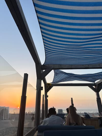 The image depicts a sunset view from a covered outdoor seating area at a beachside establishment in Hook of Holland, Netherlands. The sky is a soft gradient of blue and orange, with the sun low on the horizon. The seating area is partially enclosed by glass panels and features a striped blue and white fabric canopy supported by wooden beams. In the foreground, two individuals are seated at a table, facing away from the camera and towards the beach and sea. A man in a light-colored shirt is on the left, and a woman with long blonde hair is on the right. The beach is visible with stacks of lounge chairs and several people walking. The sea stretches to the horizon.