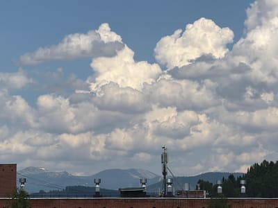 This is a wide-angle, eye-level, outdoor shot of a cityscape taken from a rooftop. The sky is filled with dramatic, fluffy white clouds against a bright blue backdrop. Below the clouds, a line of mountains forms the distant horizon, with some areas appearing snow-capped. In the middle ground, a dense forest stretches across the landscape. In the foreground, a brick building with several silver ventilation pipes and a telecommunications tower is visible. The image is calm and serene, with no people or animals present. The overall impression is one of natural beauty and urban infrastructure coexisting.