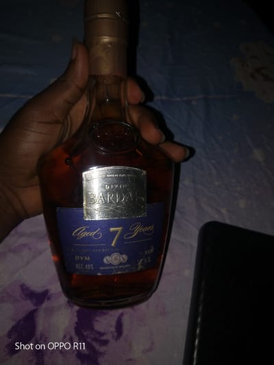 This is a close-up, low-light image of a hand holding a bottle of Divin Bardar aged 7 Years cognac. The bottle is made of clear glass and contains a dark amber liquid. The label is predominantly dark blue with silver and gold text. Visible text on the label includes "DIVIN BARDAR," "Aged 7 Years," "DVM," "ALC.40%," "PRODUCED IN MOLDOVA," and "XO 0.51." There are also embossed details on the bottle itself, including "1929." The hand holding the bottle has dark skin and painted fingernails. The background appears to be a patterned fabric, possibly bedding, in shades of blue and purple, with some white and pinkish markings. A black object, likely a phone, is visible in the lower right corner of the frame, casting a shadow. The bottom left of the image shows a watermark that reads "Shot on OPPO R11." The lighting is dim, suggesting the photo was taken indoors at night or in a poorly lit environment. The overall impression is casual, perhaps a personal moment captured while showcasing the beverage.