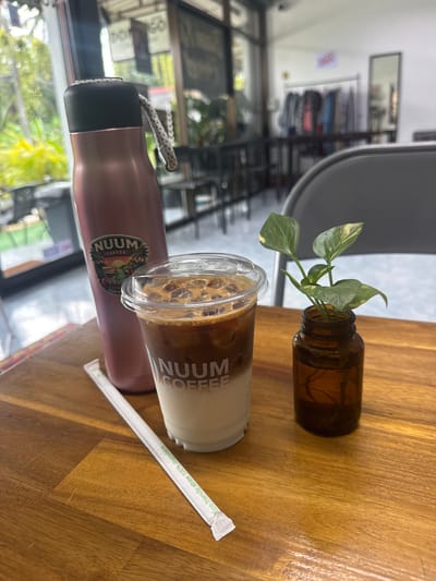 On a wooden table, located indoors in Baan Kai, Thailand, is a collection of items. A small brown glass jar holds three green and yellow pothos plants. Next to it is a clear plastic cup filled with a layered beverage, with the words "NUUM COFFEE KOH PHANGAN" visible on the side. Adjacent to the cup is a sealed white paper straw with green text. A pink metal water bottle is partially visible on the left side of the table, with the "NUUM -COFFEE-" logo. The background shows a clothing store with clothes hanging on racks, visible through a large window. A gray chair is partially visible in the upper left corner.