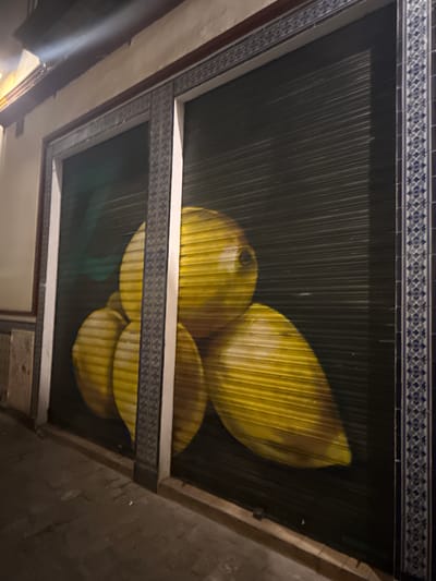The image depicts a mural painted on a dark, corrugated metal roll-up door of a storefront. The mural features a cluster of three large, yellow lemons, with a subtle green leaf visible behind them. The lemons are rendered with a high degree of realism, showcasing their textured skin and glossy surfaces. The door itself is framed by decorative tiles with a blue and white geometric pattern. The surrounding building facade is light-colored, and the ground in front of the storefront is paved. The lighting suggests it is nighttime or a dimly lit area. This mural is located in the Santa Cruz neighborhood of Seville, Spain, a historic district known for its narrow, winding streets and charming architecture.