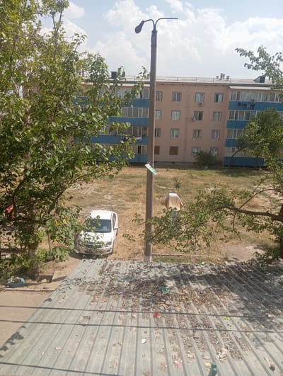 The image shows an outdoor scene in Nurabad, Uzbekistan. The view is from a higher vantage point, looking down on a courtyard area between apartment buildings. The sky is blue with scattered white clouds, suggesting it is daytime, likely late morning or early afternoon.
In the foreground, to the left, a large tree with green leaves partially obscures the view. To the right, a corrugated metal roof is visible, covered in debris, including leaves and small pieces of trash. A streetlamp pole stands vertically in the middle of the frame.
Behind the trees and the pole, apartment buildings are visible. The building on the left is multi-story with a light pink facade and blue accents around the windows. Air conditioning units are mounted on the exterior walls. Further back, another apartment building with a light blue facade is partially visible.
On the ground, between the buildings and under the trees, is a patch of dry, overgrown grass. A white car is parked on the grass. A person, partially obscured by the car and greenery, appears to be bending down near the car, possibly working on it or retrieving something. They are wearing dark clothing.
A small green and white sign is attached to the streetlamp pole. The overall impression is a residential area in a somewhat unkempt state, with evidence of human activity. There is no discernible text other than on the small sign.