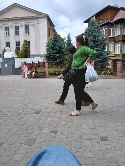 Two women are walking across a cobblestone plaza in Truskavets, Ukraine. The woman in the foreground is wearing a green, polka-dot shirt and black pants, carrying a white plastic bag in her right hand. The woman behind her is wearing a brown long-sleeved shirt and black pants. In the background, a statue of a man in a suit stands in front of a light-colored building with columns. To the right, a wooden building with balconies has a sign with Cyrillic script. Other people are visible sitting on a bench and standing near the statue. Trees, including several tall evergreen trees, are interspersed throughout the plaza. The sky is overcast.