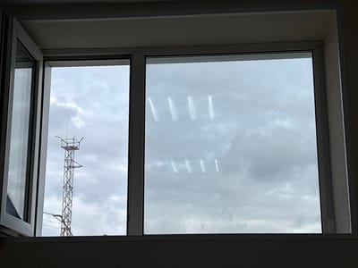 The image shows a view through a window onto an overcast sky. The window is divided into two sections. The left section is open and reveals a tall, metal lattice tower with several platforms and lights at the top, suggesting it could be a telecommunications or observation tower. The sky is filled with dense, gray clouds, indicating a dull or rainy day. The right section of the window has several smudges or streaks on the glass, appearing as faint white vertical lines. These marks could be water droplets or some form of dirt. The overall scene is likely an outdoor environment in a populated area, given the presence of the tower. The muted colors and cloudy sky create a somber or neutral mood. There is no visible text in the image.