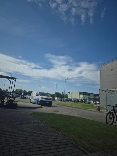 The image depicts a street scene in Pabradė, Lithuania, on a clear day with scattered clouds. In the foreground, a paved walkway leads towards a grassy area. To the left, a covered structure with metal supports is visible, and next to it, a red motorized scooter with a basket is parked. A white van with blue and orange decals, displaying the license plate MYY 807, is parked on the right side of the road. Across the road, a line of parked cars is visible in front of a large, modern building with a light-colored facade. Further in the background, additional buildings and trees can be seen. On the far right of the image, a bicycle is parked next to a covered bike rack. The overall environment suggests an outdoor commercial or industrial area.
