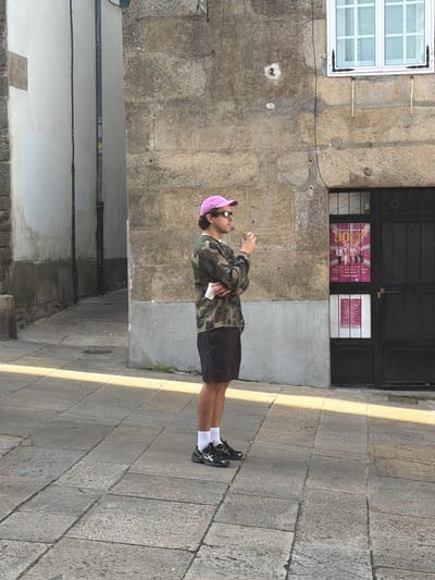 The image depicts a young person standing on a cobblestone street. The person is wearing a camouflage jacket, black shorts, white socks, and black sneakers. They are also wearing sunglasses and a pink baseball cap. The person is holding a white object, possibly a phone or a cup, in their right hand.
The background features a stone building with a large black door and a window with posters. To the left of the door, there is a white window with multiple panes. The street is paved with large, irregular granite stones. Sunlight casts long shadows across the pavement, suggesting it is either morning or late afternoon. The scene appears to be in a European city, consistent with the given location context of Santiago De Compostela, Spain. The overall mood of the image is casual and relaxed.