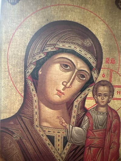 The image is a close-up of an icon, likely painted on wood, depicting the Virgin Mary and the Christ Child. The figures are rendered in a traditional Byzantine style with large, expressive eyes and somber expressions. The Virgin Mary is on the left, wearing a deep red veil and robe, with a golden halo. Her left hand rests on the shoulder of the Christ Child, who is on the right, facing forward and wearing a red robe. He also has a golden halo. Both figures have red halos with golden accents. The background is entirely gold leaf, which is characteristic of icons. There are some red Greek letters visible on the background, to the right of the Virgin Mary's head. The overall condition of the icon appears aged, with visible craquelure on the surface. The location in Marten, Bulgaria, is not depicted in this image.