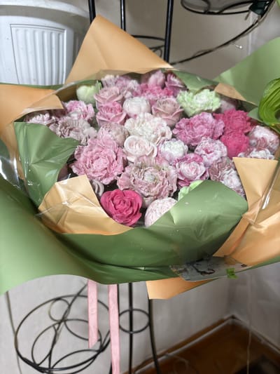 A bouquet of flowers is displayed in a close-up, eye-level shot. The bouquet features a variety of pink and white carnations, along with a few light green blossoms. The flowers are intricately arranged and appear to be made of soap or a similar material. The bouquet is wrapped in green and tan paper, with clear plastic over the flowers. Two pink ribbons hang from the bottom of the bouquet. The background is out of focus, showing a white textured surface on the left and a blurred, dark metal stand on which the bouquet is resting. The overall impression is of a decorative arrangement. The image location is Sibir, Russia.