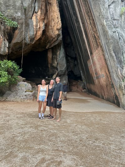 Three individuals, two females and one male, stand in front of a large cave entrance. The female on the left wears a white tank top and blue shorts. The female in the middle wears a black tank top and a dark skirt. The male on the right wears a black t-shirt and dark shorts. They are positioned on a sandy ground with small pebbles. Behind them, a massive rock formation features a dark cave opening. A sign with Thai script and "KHAO PHING KAN" is visible to the left of the cave. To the right of the cave entrance, the rock face displays vertical striations of various colors. Another individual, a male wearing a dark shirt and tan shorts, is visible walking further into the cave entrance in the background. The overall environment appears to be a natural, outdoor setting, likely a tourist attraction in Tambon Krasom, Thailand, known for its distinctive rock formations and caves.