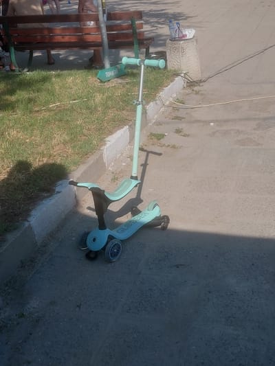 The image depicts an outdoor scene in Kostievo, Bulgaria, under bright daylight. A light blue and black Globber children's scooter, featuring a seat attachment and three wheels, is positioned on a paved surface in the foreground, casting a shadow to its right. To the left, a concrete curb separates the paved area from a narrow strip of dry grass. On this grass, partially obscured, rests a green object, possibly a small board or toy, bearing the Cyrillic inscription "ПОМАГАЙ" (meaning "HELP").
Behind the grassy strip, a wooden park bench with dark metal supports is visible. Several individuals are seated on or near the bench, with only their lower torsos and legs, clad in casual attire and footwear (including sandals), discernable. To the right of the bench, a concrete pedestal with a textured surface functions as a makeshift table, holding two plastic water bottles (one with a blue cap, one with a white cap) and some white cardboard food packaging. Three electrical cords, two dark and one lighter, run across the paved ground, originating near the base of the pedestal and extending out of frame to the right. Strong sunlight from the left casts prominent shadows from the scooter, people, and potential overhead obstructions onto the ground and grass.