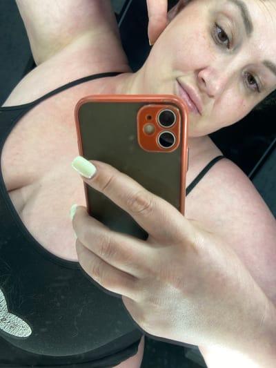 A woman is taking a selfie with her phone. She is wearing a black tank top and has light-colored nails. The background is dark and out of focus. The image is not located in Rozkvit, Ukraine.