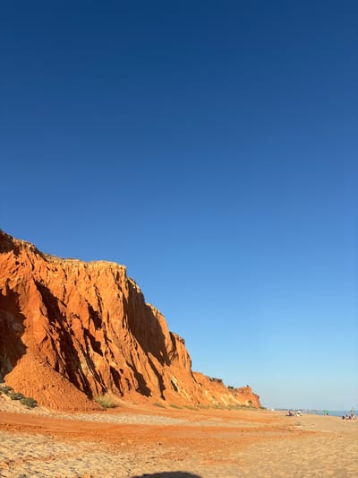 A wide, vertical shot captures the striking orange cliffs of Falesia Beach in Albufeira, Portugal, under a vast, clear blue sky. The cliffs, illuminated by the sun, dominate the left side of the frame, their textured surfaces displaying streaks of lighter sand and shadow. The foreground consists of a sandy beach, with some areas of darker, damp sand and footprints visible. In the distance, along the shoreline, several people are scattered on the beach and near the water, some with umbrellas and fishing rods, indicating a tranquil day at the seaside. The ocean is a calm, pale blue, meeting the horizon line.
