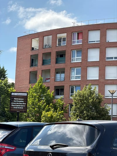 This media file captures a vertical outdoor scene dominated by a red brick apartment building with numerous windows, many featuring white roller shutters. Lush green trees with dappled sunlight are visible, partially obscuring the building and suggesting a daytime setting with good weather. A dark-colored car, likely a hatchback or SUV, occupies the right side of the frame, its reflective surface mirroring the sky and clouds.
On the lower right of the image, a sign with the JCDecaux logo is prominently displayed. The sign contains text in French, providing information about a local municipal annex. Specifically, it reads: "Informations," "Mairie annexe des Pyramides," "Espace des Deux Bois," "Place Camille Guérin," and a phone number "01 60 78 99 50." The overall impression is one of a residential urban environment in Évry-Courcouronnes, France, on a bright, clear day. The presence of scaffolding on the left side of the building indicates potential ongoing maintenance or construction.