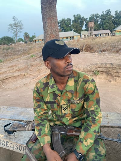 A man in a Nigerian Army camouflage uniform sits with an AK-47 across his lap. He wears a black baseball cap with a bird design. The uniform patch indicates "NIGERIAN ARMY." The man's rank insignia shows two chevrons. He also wears a watch on his wrist. The background includes a dry, open area with buildings and trees. The image has a slightly tilted perspective.