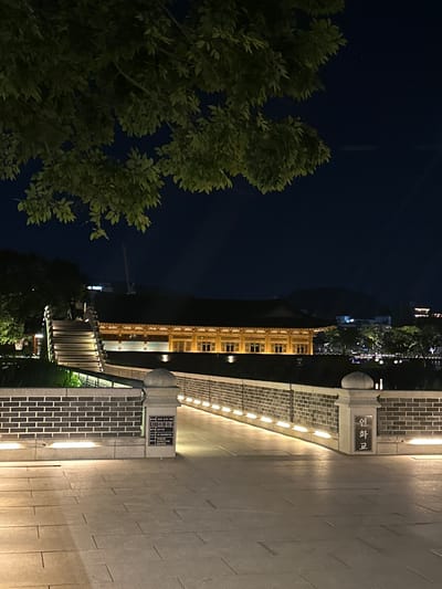 The image depicts an outdoor nighttime scene, likely in the city of Jeonju-si, South Korea, showcasing a traditional Korean architectural structure illuminated by warm lighting. In the foreground, a paved walkway made of large, square stone tiles stretches out, with subtle lines indicating the grout. To the left, a brick wall borders the walkway, with small, upward-facing lights embedded along its base, casting a glow upwards. A stone pillar with a rounded cap stands in front of the brick wall, featuring a sign with Korean text. Further back, a set of wide, illuminated stone stairs ascends to the right, with groups of people visible at the top. Beyond the stairs, a low brick wall continues, also lit from below. A prominent traditional Korean building with a dark, tiled roof and a wooden facade, adorned with multiple windows, is visible in the mid-ground. The building is brightly lit from within and by external lights. In the background, trees with dark foliage are silhouetted against the night sky. Distant city lights and the outlines of hills or mountains are faintly visible. A large tree with green leaves partially obscures the top left portion of the image. The overall atmosphere is serene and illuminated, highlighting the architectural beauty of the location at night.