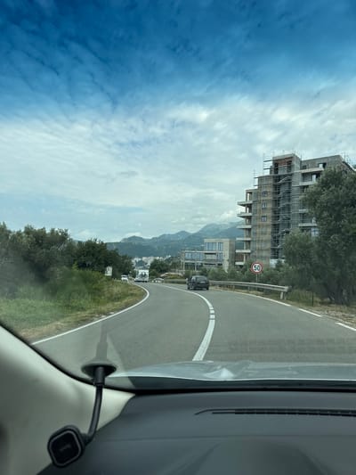The image, taken from a vehicle's interior, depicts a scenic road curving to the right in Podličak, Montenegro, during daylight hours under a partly cloudy sky. The asphalt road features clear white lane markings and a solid white line delineating its edge.
On the left side of the road, a large multi-story building is under active construction, heavily covered with metal scaffolding. The concrete structure shows multiple window openings. Behind this building, other structures and a range of mountains are visible in the distance. A red and white circular speed limit sign, displaying "50," stands prominently by the roadside near the construction site. Further ahead on the road, a dark-colored car, possibly an SUV, can be seen.
The foreground on the right reveals part of the interior of the car from which the photo was taken, including a section of the dashboard and a black charging cable or phone mount attached within the vehicle. Green vegetation, comprising trees and grass, lines the roadside, adding to the natural landscape. The sky transitions from a deep blue on the far left to a lighter, cloud-dappled expanse above the mountains.
The only clearly legible text is the number "50" on the speed limit sign. The overall scene suggests a journey through a developing coastal or mountainous region.
